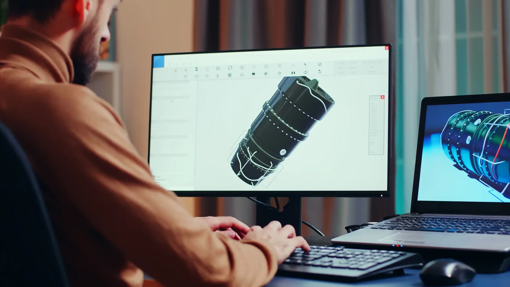 An engineer at a desk builds a 3D CAD model of a cylindrical engine component with a wiring harness on a large monitor, while a laptop beside it shows a rendered view of the same part