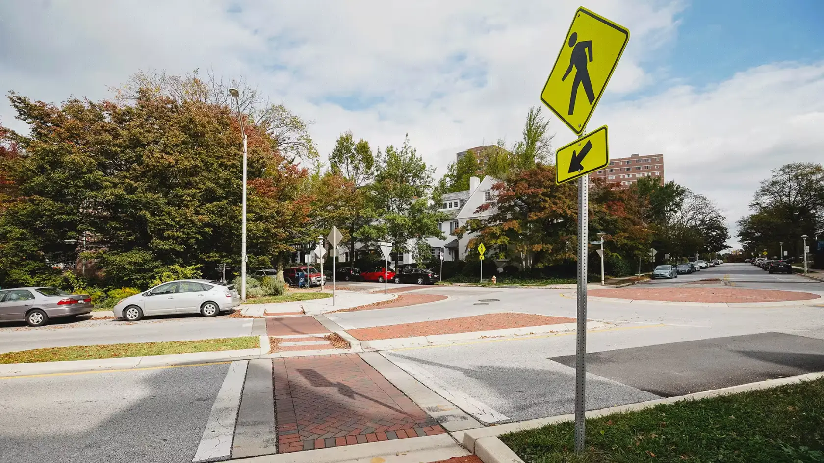 A residential intersection with a marked crosswalk, a brick pedestrian refuge island and curb ramps, under a yellow pedestrian crossing sign
