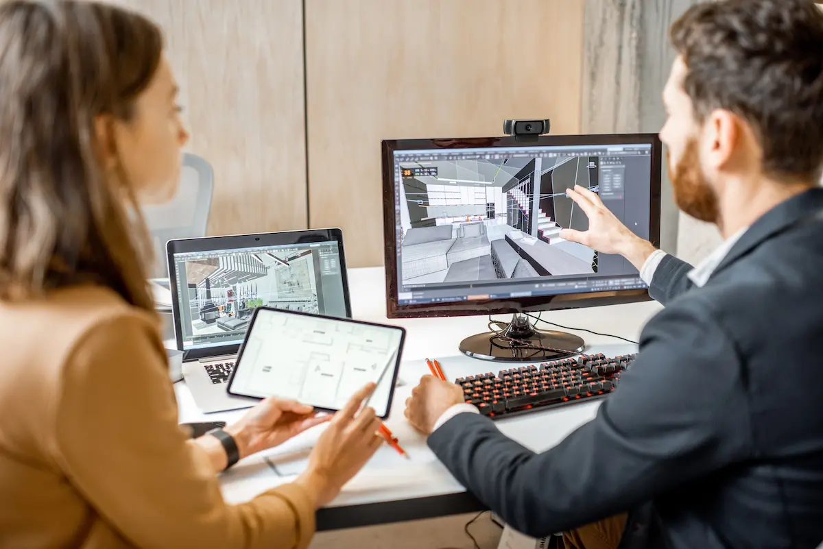 Two designers at a desk review CAD work, one pointing at a 3D building model on a monitor while the other holds a tablet showing a floor plan