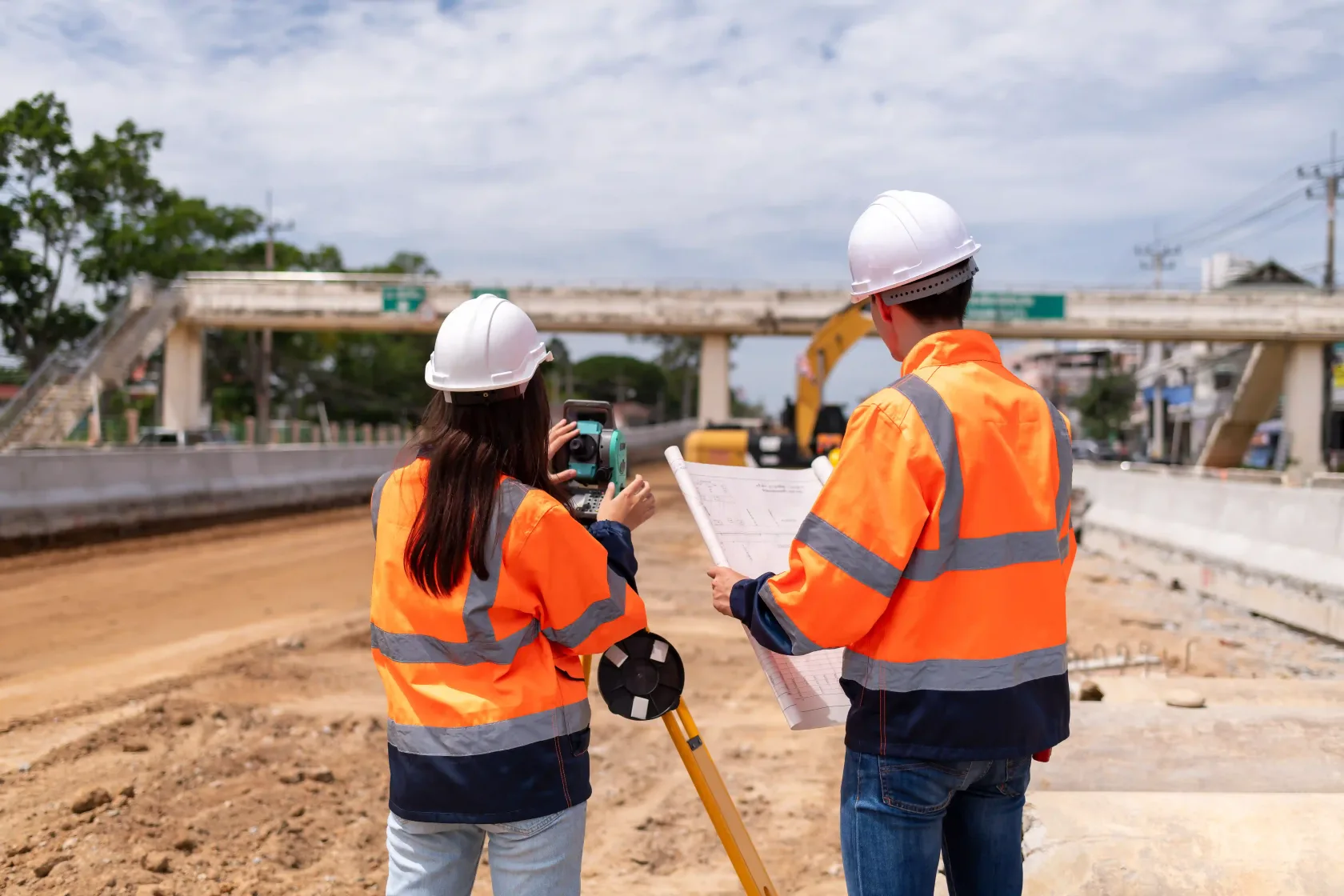 Two engineers in hard hats and high-visibility jackets on a roadway construction site, one sighting through a total station on a tripod while the other holds the plans, with a footbridge and an excavator ahead