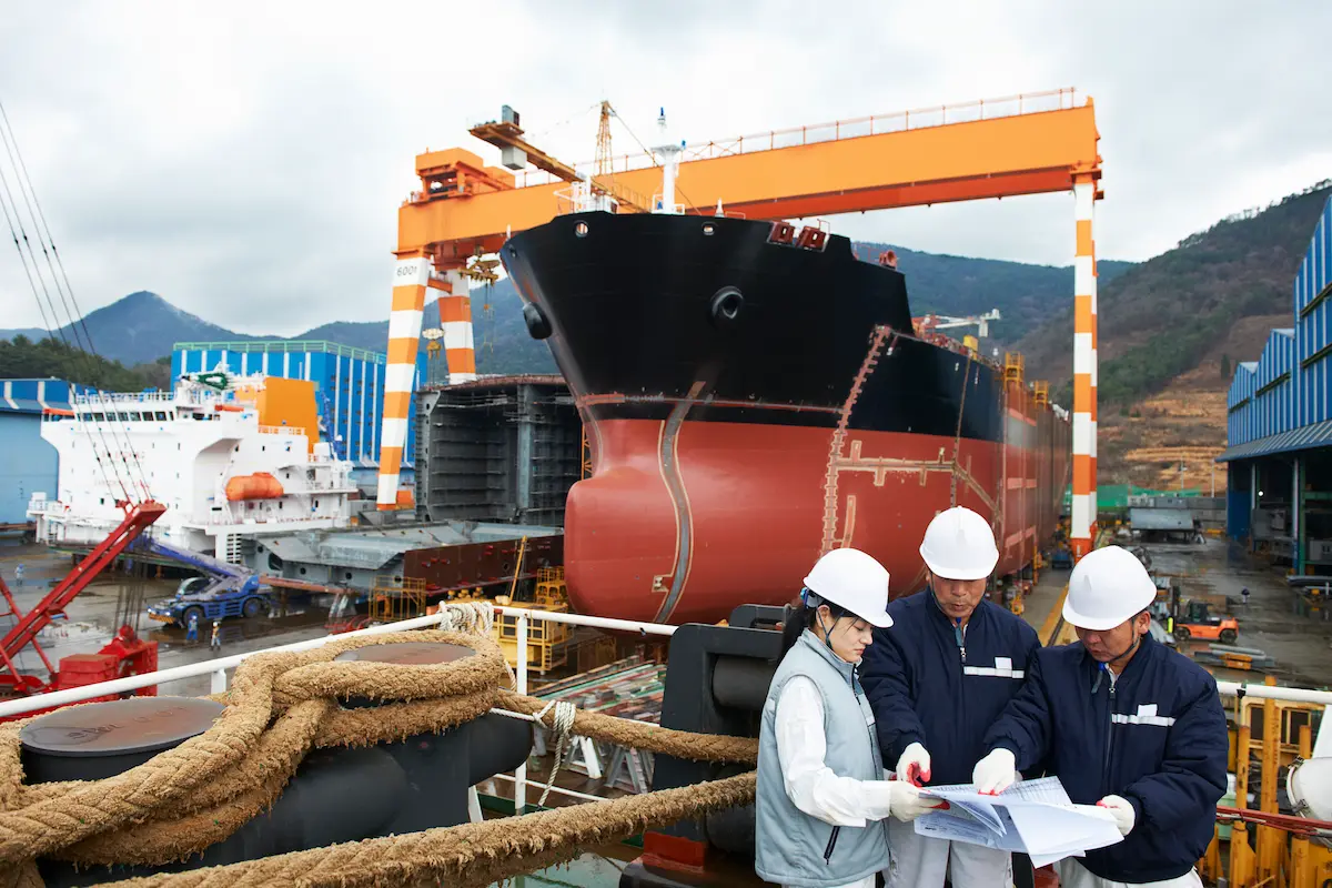 Three engineers in white hard hats review drawings on a ship’s deck in a shipyard, beside mooring ropes, while a tanker’s red and black bow stands under an orange gantry crane behind them